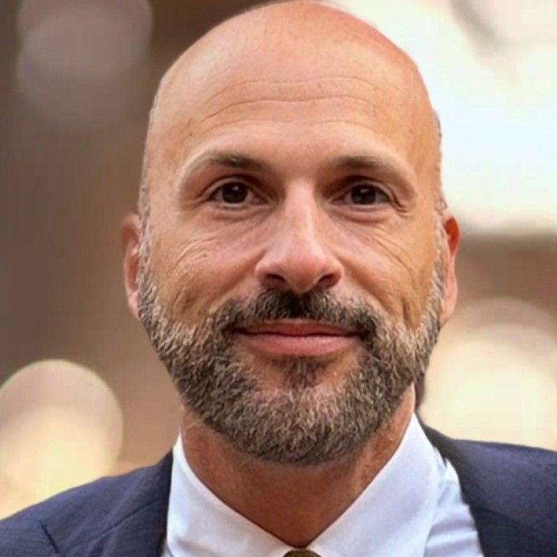 A man wearing a suit and tie standing against a blurred background.