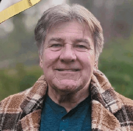 Man smiling outdoors wearing a plaid jacket and blue shirt with greenery in the background.