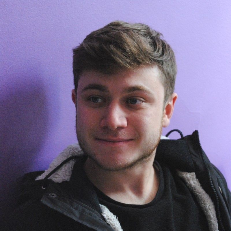 Smiling person in a black jacket sitting against a purple wall.