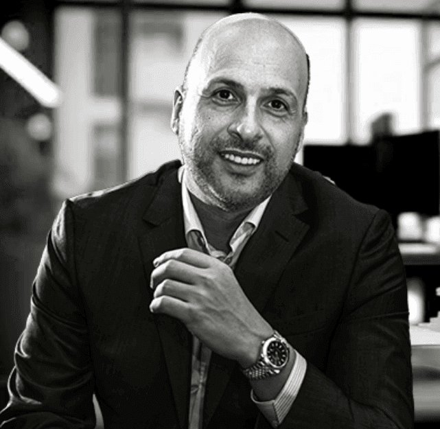 Man wearing a suit and wristwatch, sitting in an office with large windows in the background.